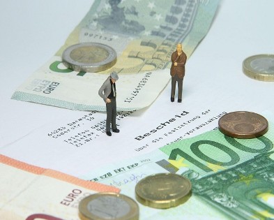 Zwei Miniaturmännchen die auf einem Papier stehen auf dem "Bescheid" steht und mehrere Geldscheine und Münzen um sie herum.